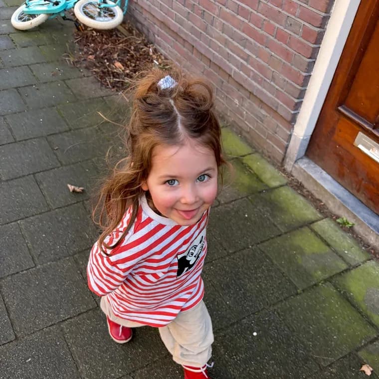 Foto van een jong kind met een staartje in een rood-wit gestreept shirt, dat omhoog kijkt naar de camera